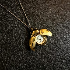 Peut inclure: Un pendentif montre en forme de coccinelle dorée sur une fine chaîne argentée. La coccinelle possède une carapace articulée qui s'ouvre pour révéler un cadran blanc rond avec des chiffres et des aiguilles noirs. L'extérieur est doré mat.