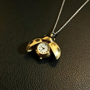 May include: A gold-toned ladybug pendant watch on a silver-colored chain. The ladybug wings are open to reveal a round white watch face with black Roman numerals and hands. The pendant features dark circular spots on the metallic gold wings.
