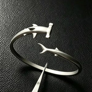 May include: A silver-toned open cuff bracelet featuring two hammerhead shark figures at the ends. The bracelet has a smooth, matte finish and a curved design. The shark heads face each other, creating a gap in the center of the circular metal band.