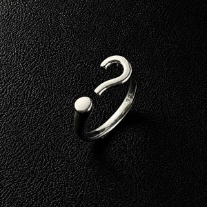 Op de afbeelding: Een zilverkleurige ring met een vraagtekenontwerp. De ring is open, met een cirkelvormige stip aan het ene uiteinde en een gebogen haakvorm aan het andere uiteinde. Het metaal heeft een gepolijste, reflecterende afwerking en een kleine stempel.