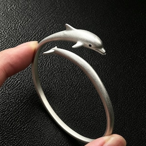 Puede incluir: Una pulsera abierta de color plateado con un motivo de delfín en un extremo. El metal tiene una textura mate y cepillada en toda la pieza, y el delfín presenta un pequeño detalle ocular circular y oscuro. La pulsera es de forma circular.