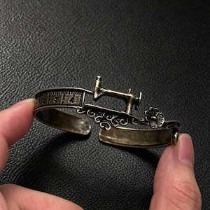 Può includere: Un bracciale a fascia in ottone anticato con un motivo in rilievo di macchina da cucire, un fiore decorativo e una banda incisa con un design a righello che mostra i numeri da uno a quattro. Il bracciale aperto in metallo ha una finitura scura.