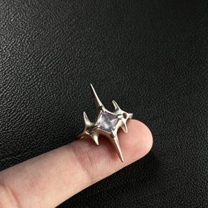 May include: A silver-toned ring featuring a four-pointed star design with elongated, sharp spikes extending from the center. A square-cut, pale purple gemstone is set in the middle of the star, creating a distinct contrast with the metallic finish.