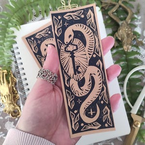 May include: Two rectangular bookmarks with a black and tan design featuring a snake coiled around a key. The bookmarks have a decorative border with leaves and geometric shapes. The design is a linocut style. A silver ring is visible.
