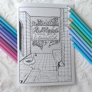 Peut inclure: Un livre de coloriage en noir et blanc intitulé "The Pool Rooms" par La Greenwitch, représentant une scène de salle de bain avec un flamant rose et une enseigne. Des crayons de couleur dans les tons de bleu et de violet sont disposés autour du livre.