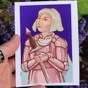 May include: A hand-painted portrait on a white card depicting a figure in pink armour holding a sword. The figure has short, light blonde hair and a serious expression. The background is a deep purple, with a light blue cape. The artist's initials are in the bottom right.