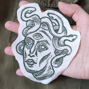 Peut inclure: Patch en tissu vert et blanc avec une illustration détaillée de Méduse, une figure mythologique grecque aux cheveux de serpents.