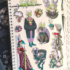 May include: A collection of colorful stickers arranged on a spiral-bound notebook page. The stickers feature various illustrations including a sword with candles, a masked figure, a winged creature, a skeleton holding a fork, and floral motifs.