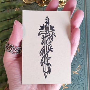 May include: A black and white illustration of a sword entwined with vines and flowers, printed on a beige card. The sword has a detailed hilt and blade. The card is held in a hand, with a decorative ring visible on a finger.
