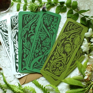May include: A collection of botanical-themed bookmarks in shades of green and white. The bookmarks feature intricate linocut designs of leaves, berries, and floral elements. The bookmarks are arranged on a lace surface, with a brass frog figurine and sprigs of greenery.