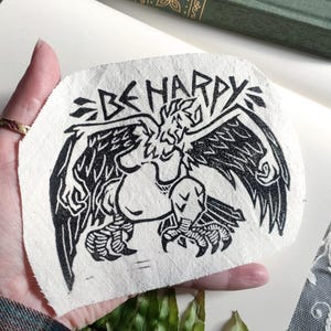 Può includere: Una toppa in tessuto bianco e nero con la scritta "BE HAPPY" sopra un'illustrazione stilizzata di un grifone. Il grifone ha ali spiegate e zampe artigliate dettagliate. La toppa è tenuta su uno sfondo bianco.