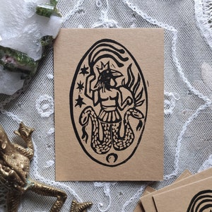 Puede incluir: Una impresión de linóleo en blanco y negro sobre papel marrón que presenta una imagen estilizada de una figura coronada con cabeza de pájaro y una serpiente envuelta alrededor de su cuerpo. La figura está rodeada de estrellas y una luna creciente.