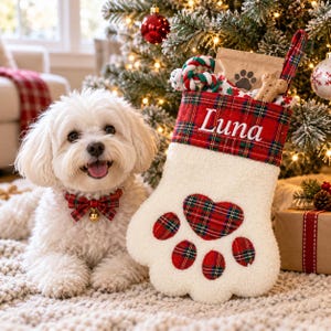 May include: A white, fluffy, paw-shaped Christmas stocking featuring a red and green plaid cuff embroidered with the name Luna. The stocking has a matching plaid heart and paw pad accents. A small white dog wears a matching plaid bow tie.