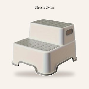 May include: A two-step stool in white and grey with the text Simply Sylka above it. The stool features a rectangular design with grey non-slip textured surfaces on both steps and integrated side handles for portability and ease of use.
