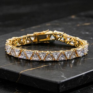 May include: A gold-toned tennis bracelet featuring a continuous row of triangular-cut clear white stones set in a metal frame. The bracelet includes a secure box clasp mechanism and is designed with a repeating geometric pattern of faceted gems.