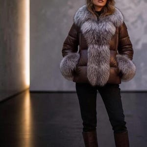Può includere: Un piumino marrone caratterizzato da un ampio colletto in pelliccia grigia soffice e polsini in pelliccia abbinati sulle maniche. La giacca è indossata con pantaloni neri e stivali in pelle marrone al ginocchio, creando un insieme coordinato.