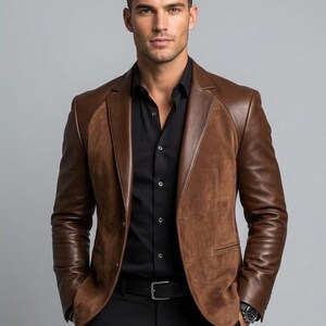 Op de afbeelding: Een bruine blazer met gladde leren mouwen en een voorpand van suède, gedragen over een zwart overhemd en een zwarte broek met een zwarte riem. De blazer heeft revers en een sluiting met twee knopen. De letters IFW zijn in de hoek zichtbaar.