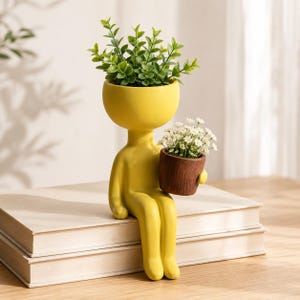 Op de afbeelding: Een gele keramische plantenbak in de vorm van een zittend figuur met een rond hoofd dat een groene bladplant bevat. Het figuur houdt een kleine bruine cilindrische pot vast gevuld met kleine witte bloemen als decoratie.
