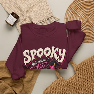 Peut inclure: Un sweat-shirt à manches longues bordeaux avec une impression graphique portant le texte "SPOOKY but make it cute" en lettres blanches et roses. Le motif comprend un chapeau de sorcière noir avec un nœud rose, une petite chauve-souris et une lune.