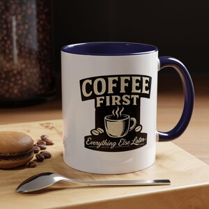 May include: A white ceramic mug with a dark blue interior and handle. The front features a black graphic design with the text COFFEE FIRST and Everything Else Later, accompanied by an illustration of a steaming cup and coffee beans.