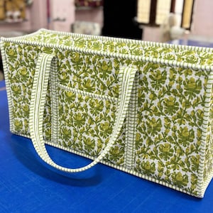 以下が含まれることがあります： 緑と白のフローラルなブロックプリント柄が施された長方形のトートバッグ。補強されたエッジを持つ構造的な形状で、2本の長いストライプ柄の布製ハンドルと平らな底を備えており、全体に一貫したキルティングの質感が特徴です。