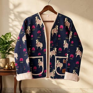 Könnte beinhalten: Eine marineblaue Steppjacke mit einem sich wiederholenden Muster aus weißen Kühen mit roten Punkten und rosa Lotusblumen. Die Jacke hat lange Ärmel mit gestreiften Bündchen, eine offene Vorderseite, zwei aufgesetzte Taschen und ein beigefarbenes Futter.
