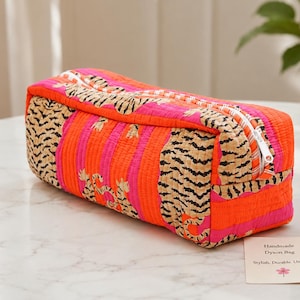 May include: A rectangular quilted zippered pouch featuring a vibrant pattern of orange and pink vertical stripes interspersed with tiger-print panels. A small rectangular tag is placed nearby with the text: Handmade Dyson Bag, Stylish, Durable, Unique.