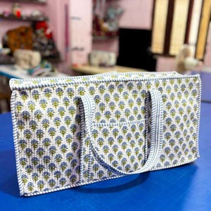 Könnte beinhalten: Eine rechteckige, gesteppte Tragetasche mit einem sich wiederholenden blau-grünen Blumenmuster auf weißem Grund. Die Tasche hat zwei passende Stoffgriffe sowie blau-weiß gestreifte Kanten an den Rändern und der Vordertasche.
