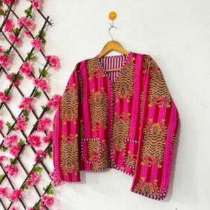Könnte beinhalten: Eine gesteppte Jacke mit einem lebendigen Muster aus orangefarbenen und schwarzen Tigern auf einem Hintergrund aus vertikalen, kräftigen rosa und dunkelroten Streifen. Die Jacke hat einen V-Ausschnitt, lange Ärmel und zwei aufgesetzte Taschen.