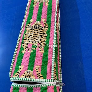 Puede incluir: Un estuche de tela acolchada largo y rectangular con un patrón repetitivo de tigres sobre un fondo de rayas verticales rosas y verdes. El estuche tiene una cremallera blanca en un extremo y un borde con rayas verdes y blancas.