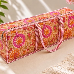 May include: A rectangular quilted fabric storage bag featuring a vibrant floral pattern in shades of hot pink, orange, and tan. The bag includes a top zip closure and two matching pink and white striped fabric handles with piped edges.
