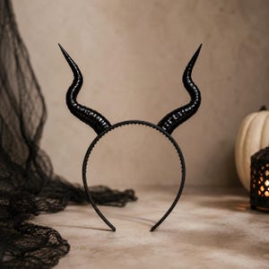 May include: A black headband featuring two large, curved, textured horns extending upward. The horns have a glossy finish with horizontal ridges along their length, and the headband itself is a thin, simple black band with a serrated inner edge.