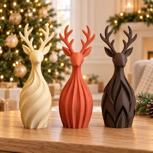 May include: Three standing deer figurines with stylized antlers. The left deer is cream, the center deer is orange, and the right deer is dark brown. Each figurine features a smooth, elongated body with a distinct vertical textured pattern.