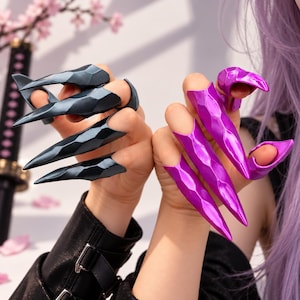 Puede incluir: Un conjunto de anillos de armadura para dedos con un diseño facetado en forma de garra. Una mano lleva cuatro anillos de color gris carbón oscuro, mientras que la otra mano lleva cuatro anillos de color púrpura magenta vibrante.