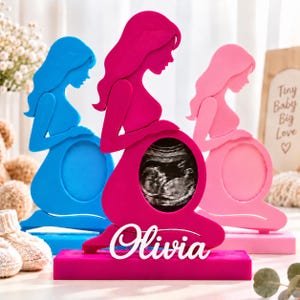 Può includere: Tre sagome in piedi di persone incinte in blu, magenta e rosa chiaro. Ogni figura presenta un ritaglio ovale centrale per un'immagine ecografica. La figura magenta ha il nome Olivia in corsivo bianco sulla base.