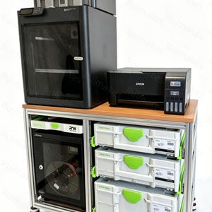 Puede incluir: Una impresora 3D negra con un sistema de múltiples bobinas acoplado se sitúa junto a una impresora Epson negra sobre una mesa de trabajo metálica. La mesa incluye un armario inferior con puerta de cristal y tres maletines grises apilados.