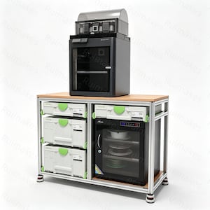 May include: A 3D printer with a clear top cover sits on a wooden workbench with an aluminium frame. The workbench includes three stacked light grey storage cases with green latches on the left and a black glass-door storage cabinet on the right.