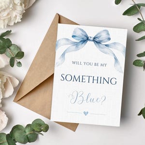 May include: A white greeting card with a watercolor blue ribbon bow at the top and the text "WILL YOU BE MY SOMETHING Blue?" in dark blue lettering, accompanied by a small blue heart icon, placed next to a brown kraft paper envelope.