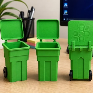 Op de afbeelding: Drie felgroene miniatuurafvalbakken met scharnierende deksels en kleine zwarte wielen. Twee van de bakken staan met de deksels open, terwijl de derde bak van achteren wordt getoond met het deksel gesloten en een cirkelvormig patroon.