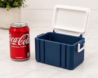 Könnte beinhalten: Eine kleine, rechteckige, marineblaue Kühlbox mit weißem Deckel und weißen Seitengriffen, die neben einer standardmäßigen 355-ml-Dose Coca-Cola steht. Die Kühlbox hat ein geripptes Außendesign und einen offenen, leeren Innenraum.