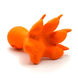 Pode incluir: Um objeto laranja brilhante impresso em 3D com uma cabeça lisa e arredondada ligada a uma base texturizada de várias pontas com cinco extensões distintas em forma de dedos. A superfície apresenta linhas de camada visíveis da fabricação.