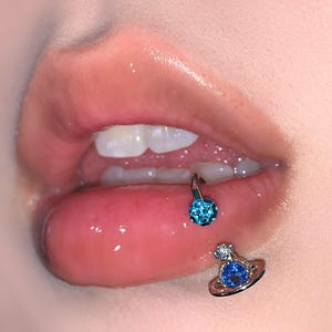 Puede incluir: Un piercing labial de tono plateado que presenta una barra circular con una bola incrustada de cristales azul brillante y un dije en forma de planeta con una piedra central azul y un pequeño cristal transparente en la parte superior.