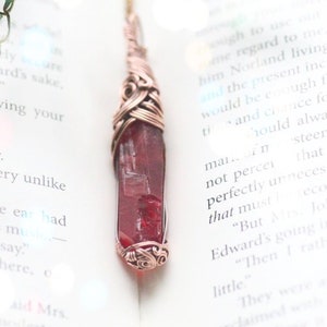 Puede incluir: Un colgante artesanal con un cristal rectangular de color rojo intenso envuelto en alambre de cobre. El trabajo de alambre tiene intrincados remolinos y patrones. El colgante cuelga de una delicada cadena, con hojas verdes en el fondo.