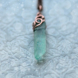 May include: A teal blue crystal pendant with a copper wire design hangs from a brown cord against a light blue background.