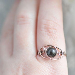 Puede incluir: Un anillo artesanal con una piedra preciosa redonda de color gris oscuro engastada en un delicado dise&ntilde;o de alambre. El metal del anillo tiene un tono c&aacute;lido, similar al cobre, y la piedra preciosa es el punto focal. El anillo se muestra en un dedo.