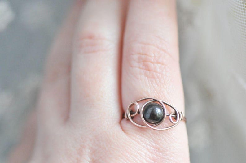 Puede incluir: Un anillo artesanal con una piedra preciosa redonda de color gris oscuro engastada en un delicado dise&ntilde;o de alambre. El metal del anillo tiene un tono c&aacute;lido, similar al cobre, y la piedra preciosa es el punto focal. El anillo se muestra en un dedo.