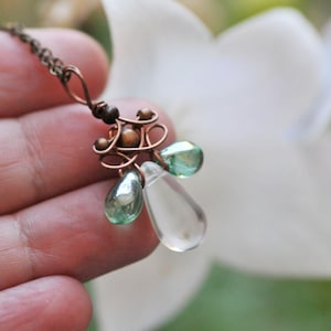 Puede incluir: Un collar de cobre hecho a mano con un colgante con cuentas verdes y transparentes en forma de lágrima. El colgante tiene un intrincado trabajo de alambre y pequeñas cuentas marrones. Fondo borroso de flor blanca.