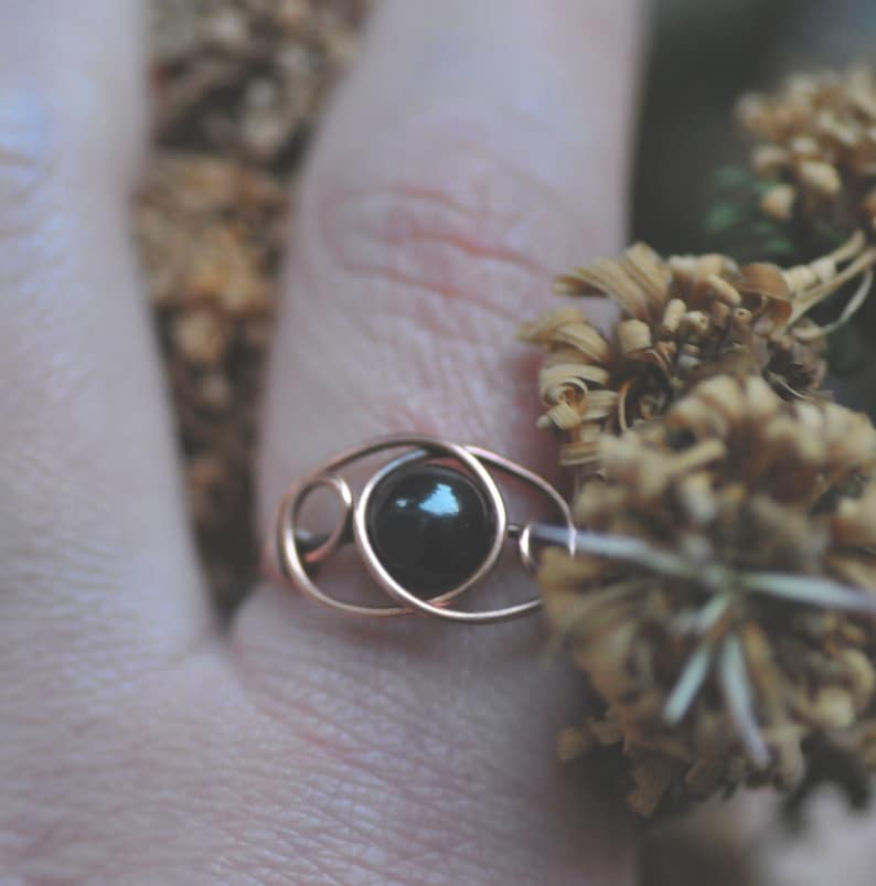 Puede incluir: Un anillo artesanal con una piedra preciosa oscura y redonda engastada en un dise&ntilde;o de alambre color cobre. El dise&ntilde;o del anillo presenta una piedra central rodeada por una serie de bucles interconectados. El anillo se muestra en un dedo, con flores secas en el fondo.