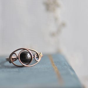 Puede incluir: Anillo artesanal de alambre de cobre con una piedra oscura y redonda en el centro. El dise&ntilde;o del anillo incorpora patrones de alambre en bucle y remolino, creando una est&eacute;tica abierta y aireada. El anillo est&aacute; sobre un fondo azul suave.