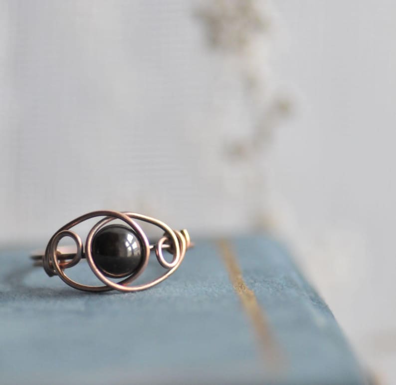 Puede incluir: Anillo artesanal de alambre de cobre con una piedra oscura y redonda en el centro. El dise&ntilde;o del anillo incorpora patrones de alambre en bucle y remolino, creando una est&eacute;tica abierta y aireada. El anillo est&aacute; sobre un fondo azul suave.